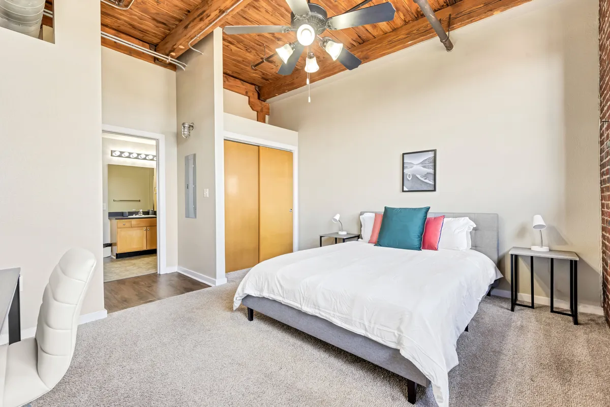 A tranquil bedroom scene with a bed, ceiling fan, nightstands, and soft artwork. Natural light brightens the space, offering a soothing setting for relaxation or reading.