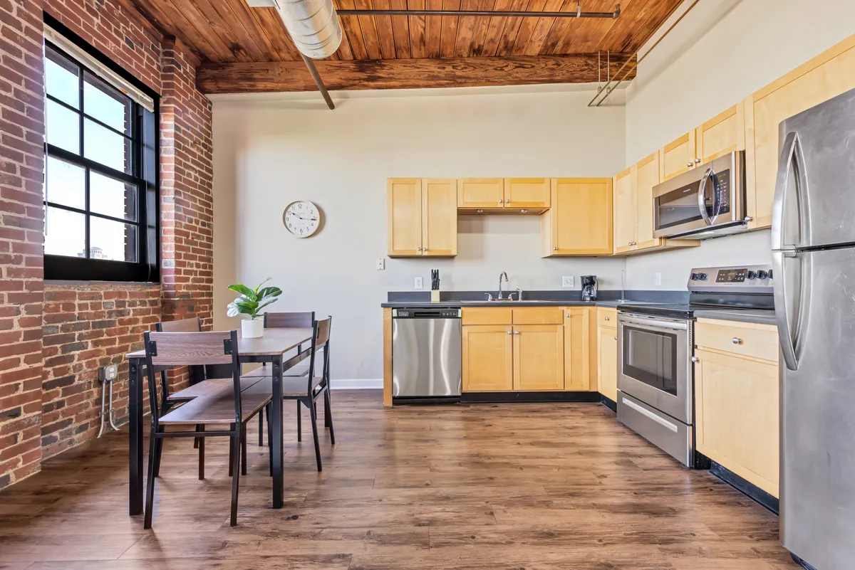 A warm, cozy kitchen and dining area with stylish cabinetry, a spacious dining table, exposed brick walls, and a large window flooding the space with natural light. The setup invites both intimate dinners and casual morning coffee.