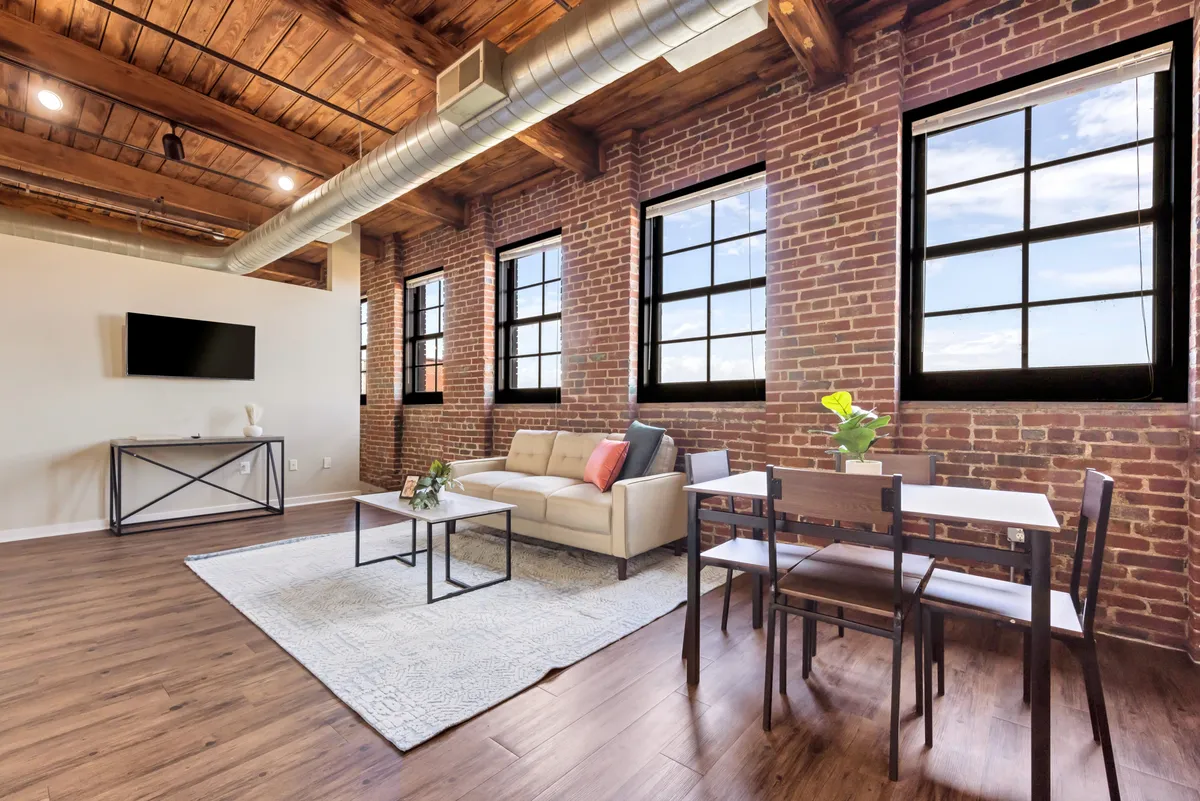 A spacious living lounge is shown with a comfortable sofa, a dining table, and oversized windows set against exposed brick walls. Natural light floods the space, highlighting both the plush modern furnishings and the original architectural elements such as hardwood floors and large windows. The ambiance is one of relaxation and sociability.