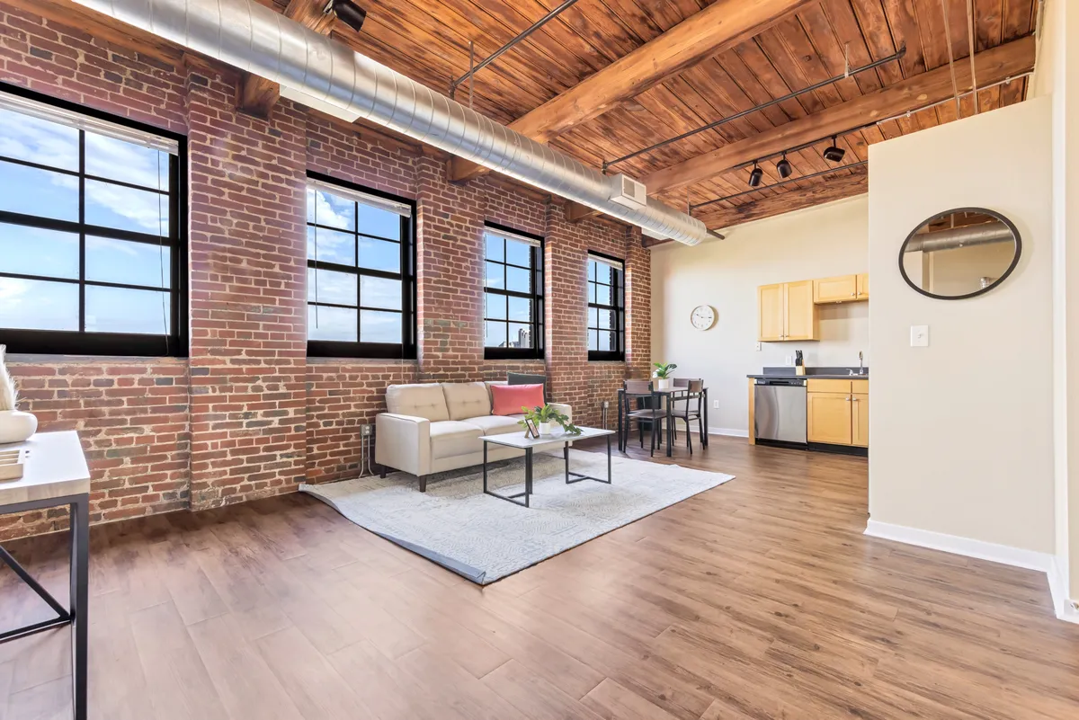 A stylish interior featuring exposed brick walls and large windows that fill the flexible nook with sunlight. The space includes a comfortable sofa seating area and a kitchenette, offering both a relaxing atmosphere and practical amenities for working, reading, or enjoying a quiet moment.