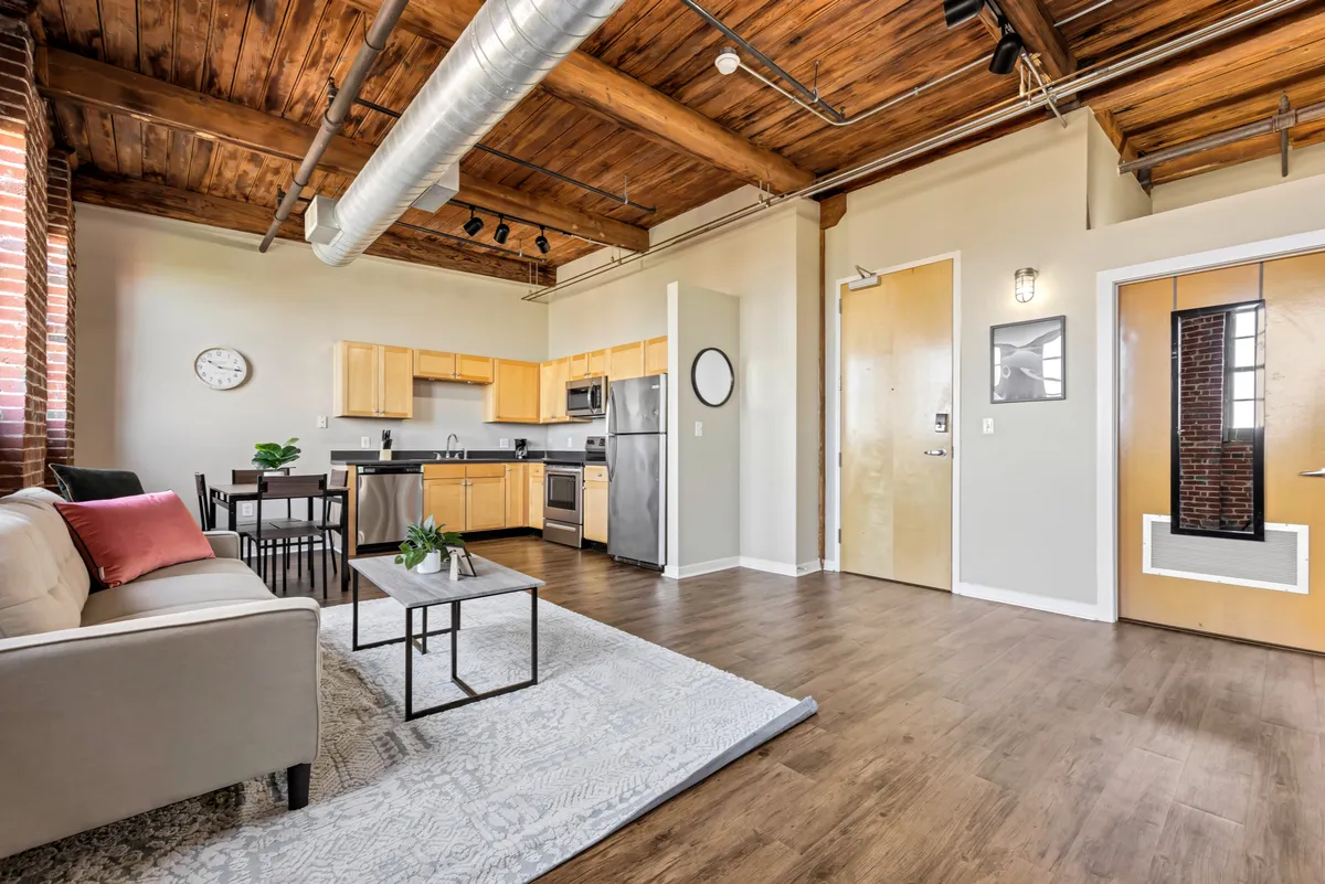 A modern, open-concept living space that seamlessly integrates the kitchen area, a dining table, and a comfortable sofa. Wooden beams, a decorative rug, and soft lighting combine with the kitchen zone to evoke both comfort and style.