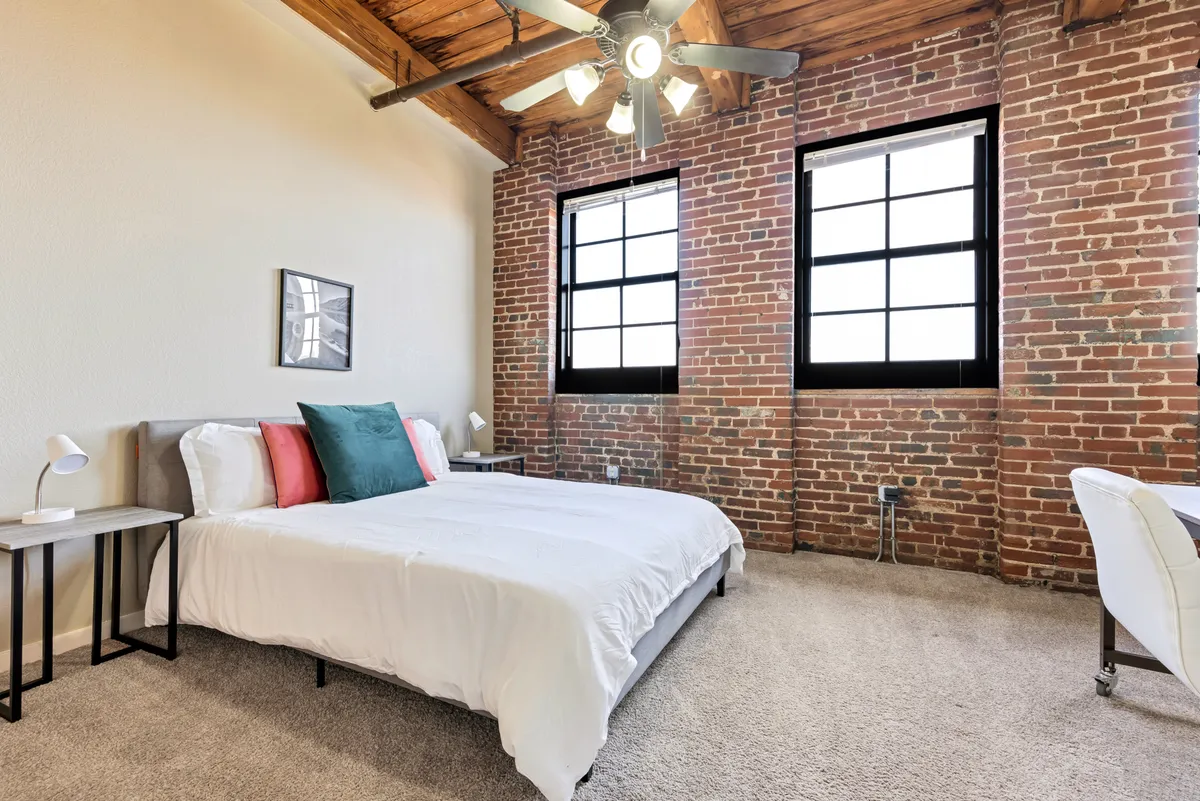 The image showcases a serene bedroom with a plush bed facing tall windows that flood the space with natural light. An exposed brick wall adds historic character, while a cozy reading nook and desk invite relaxation or quiet productivity.