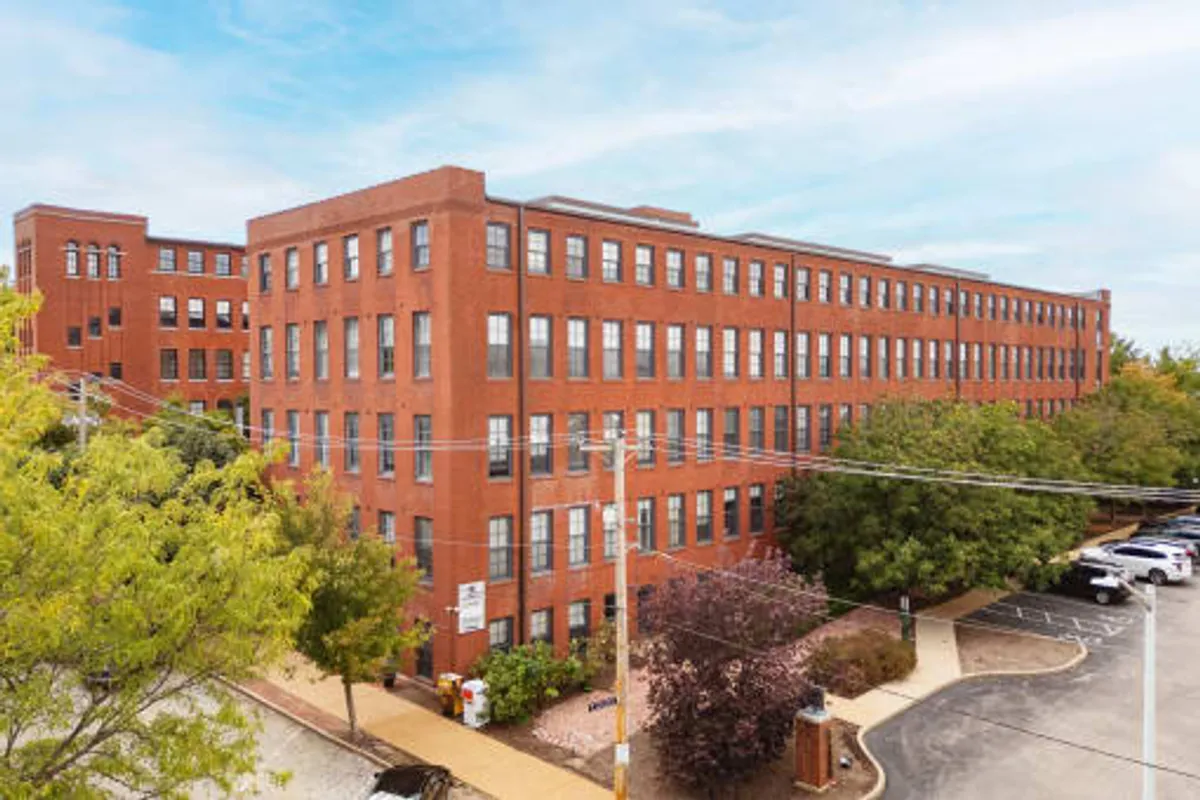 This image captures the exterior of a red brick building with large windows, surrounded by greenery and adjacent to a relaxed parking area. People can be seen walking and socializing near a nearby park, with hints of the vibrant neighborhood and historic architecture visible in the scene.