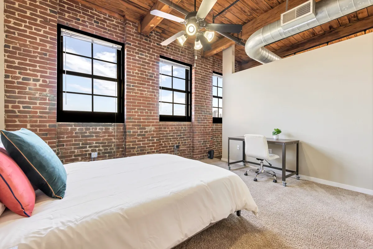 A bright and stylish bedroom featuring exposed brick walls, a comfortable bed with colorful pillows, a ceiling fan overhead, and a desk with a chair by the window. The room is filled with natural light, highlighting the unique mix of historic character and modern comfort.