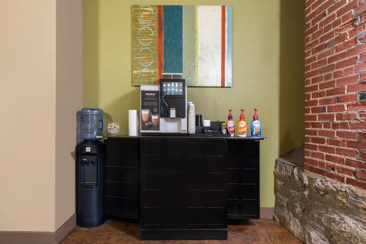 A bright, welcoming coffee corner featuring a modern coffee machine, a water cooler, and vibrant artwork on the wall. The space is inviting, encouraging guests to make themselves at home while enjoying a warm beverage or engaging in informal conversation.