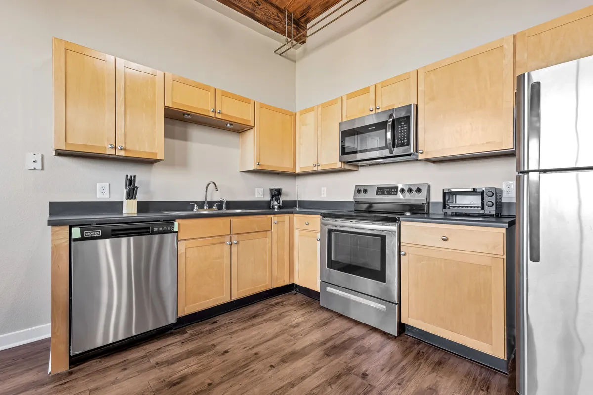 A bright, modern kitchen featuring sleek stainless steel appliances, light wood cabinetry, and a striking black countertop. The open layout and ample prep space create an inviting atmosphere for cooking, entertaining, or gathering around the kitchen island.